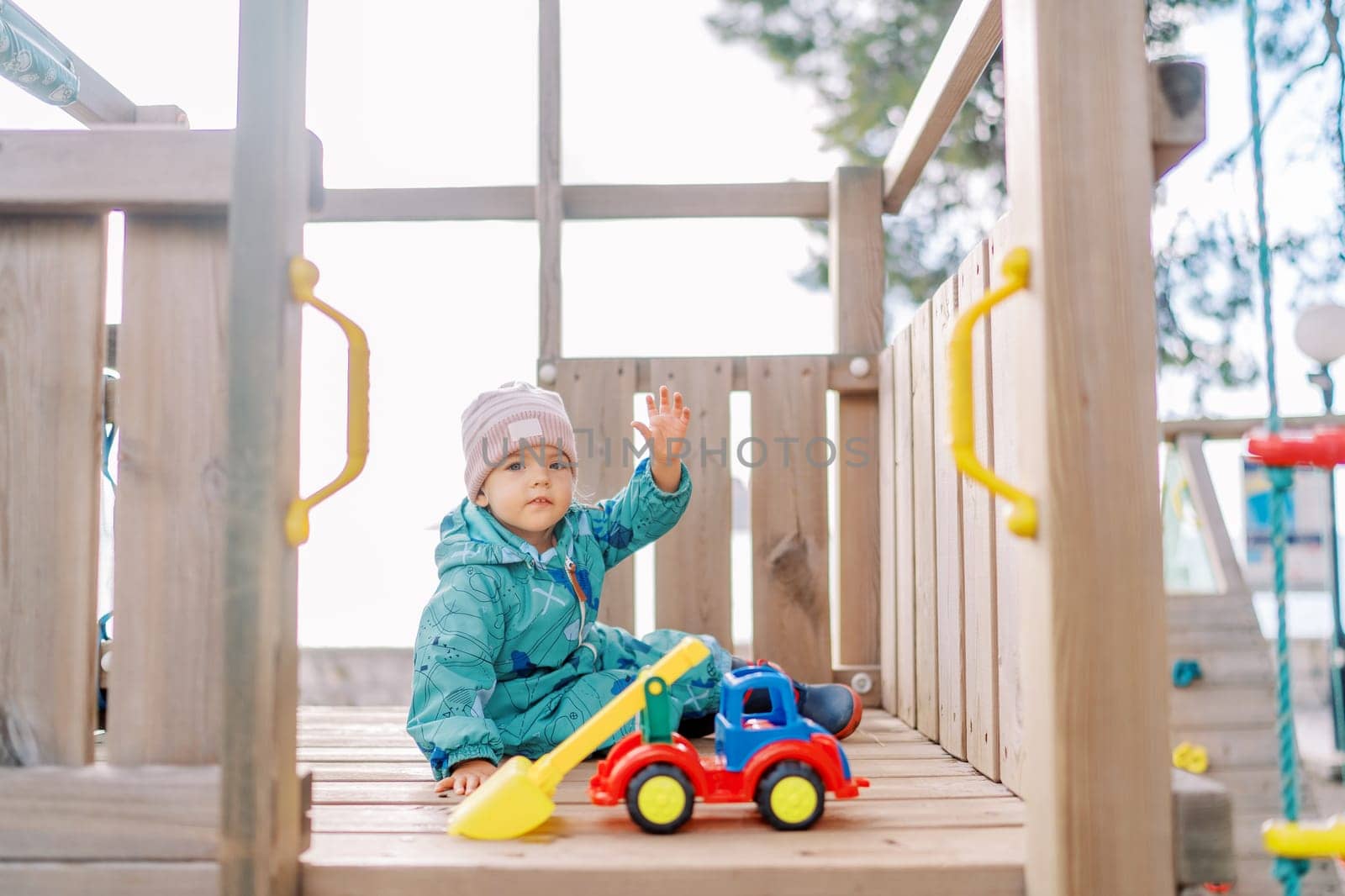 Little girl sits on a slide near a plastic toy forklift and waves her hand. High quality photo