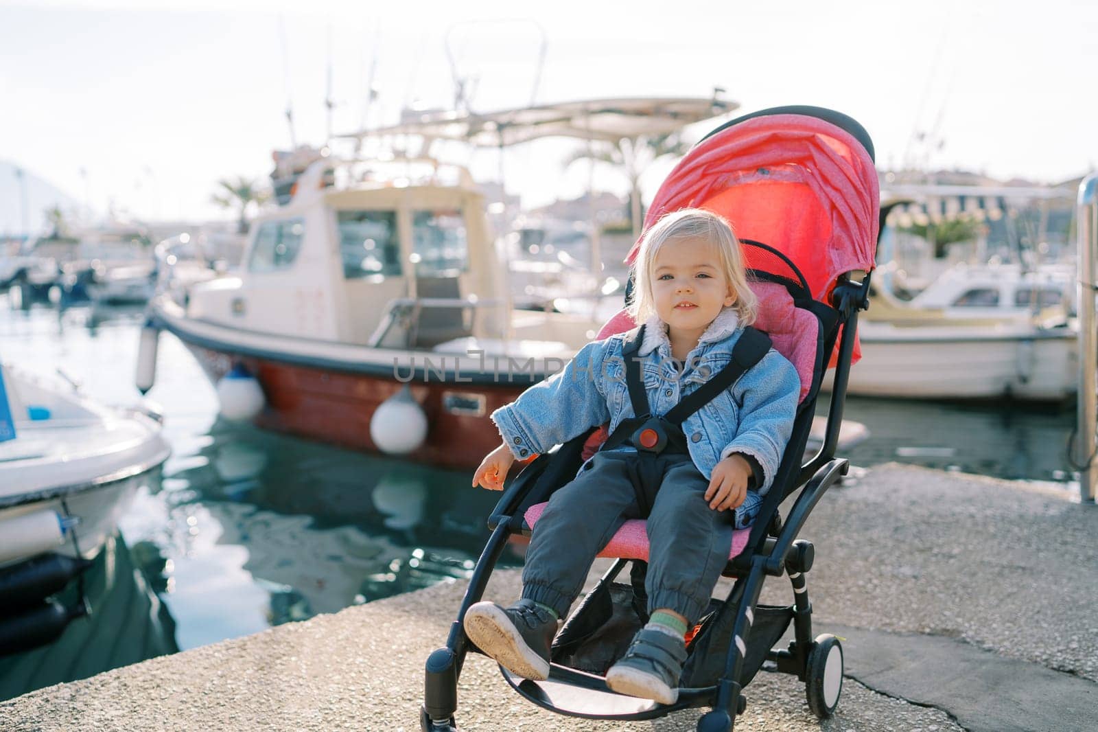 Little smiling girl sits in a stroller on the pier near the moored yachts and points to the sea. High quality photo