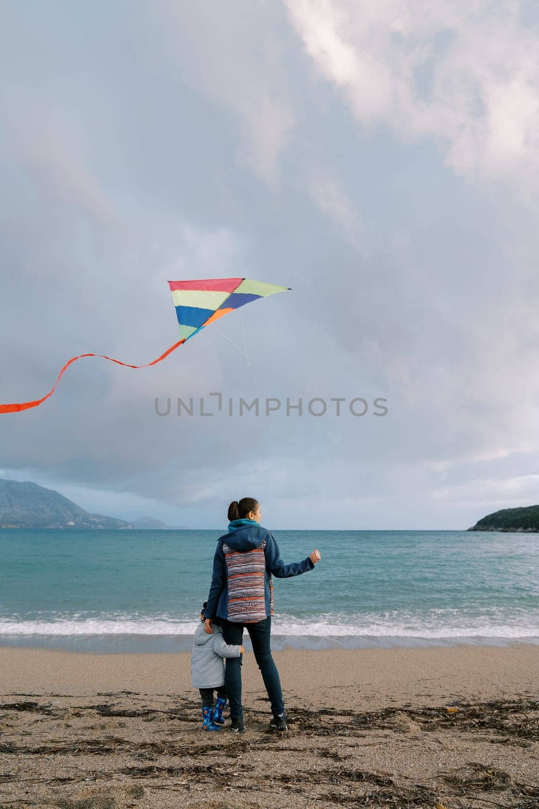 Small girl hugs her mother leg standing on the seashore with a kite. Back view by Nadtochiy