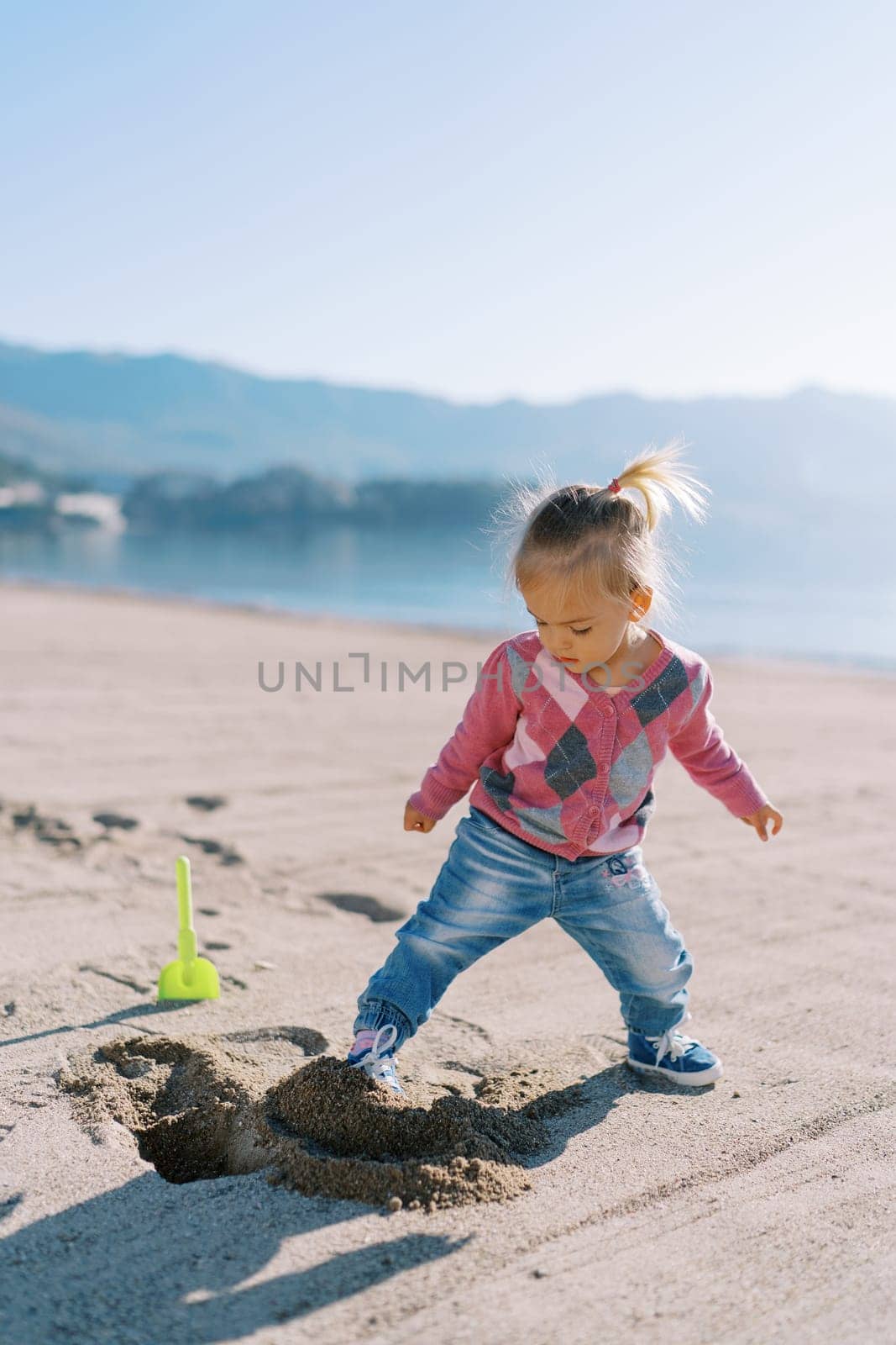 Little girl fills a hole in the sand with her foot near a toy shovel. High quality photo