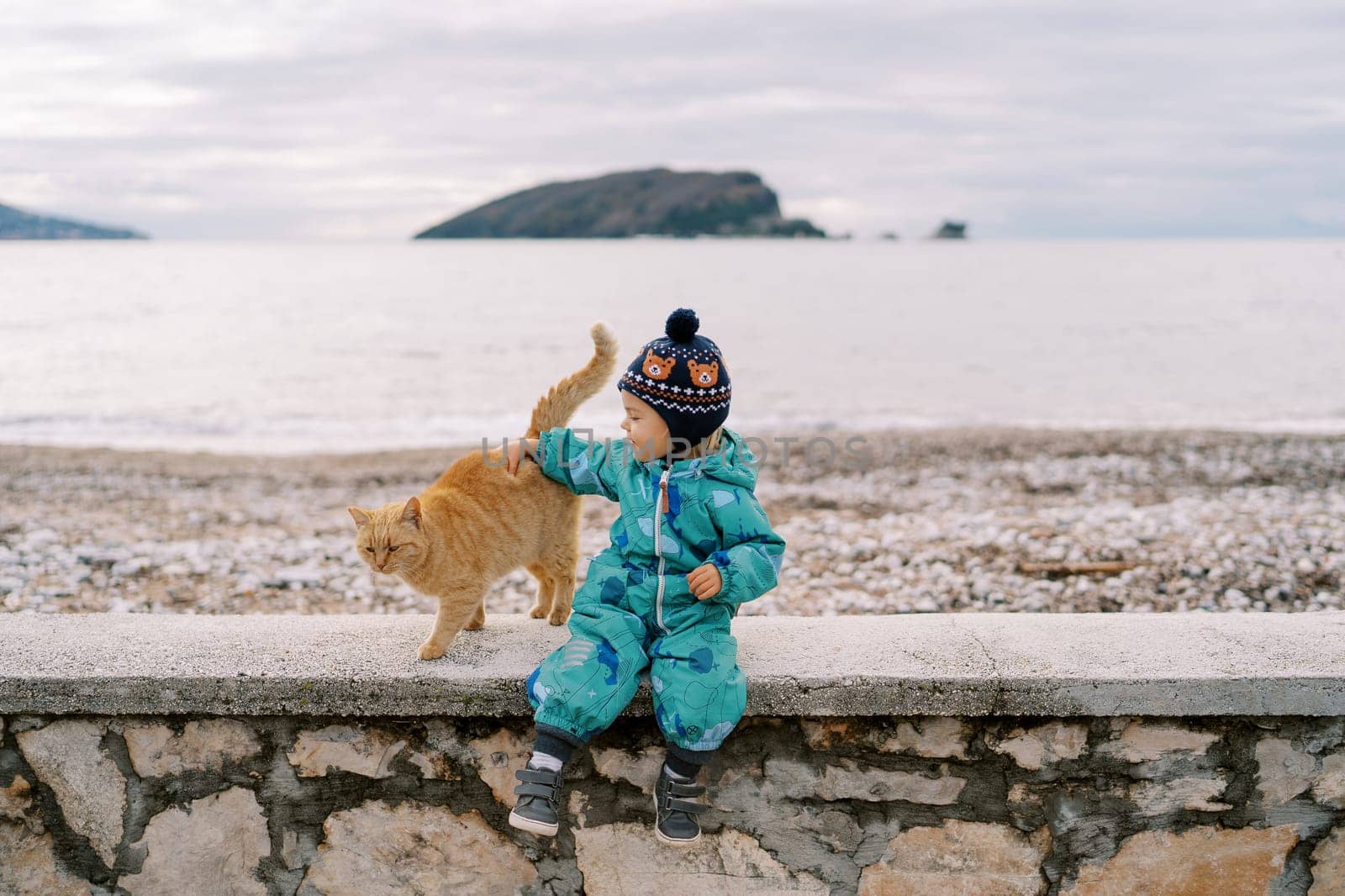Little smiling girl petting a ginger cat sitting on a stone fence by the sea by Nadtochiy