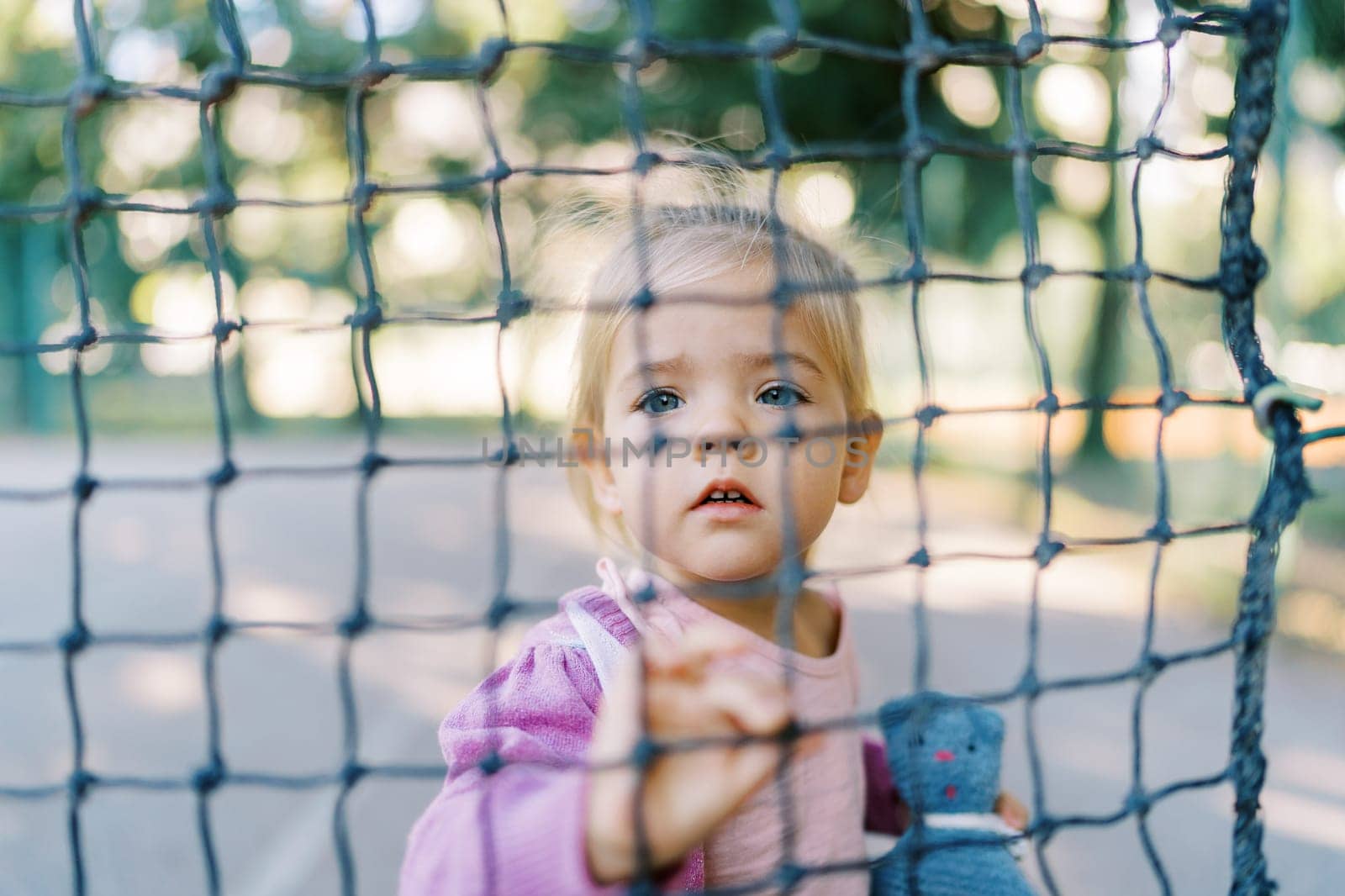 Little girl with a toy stands behind a tennis net, holding a cell with her hand. High quality photo