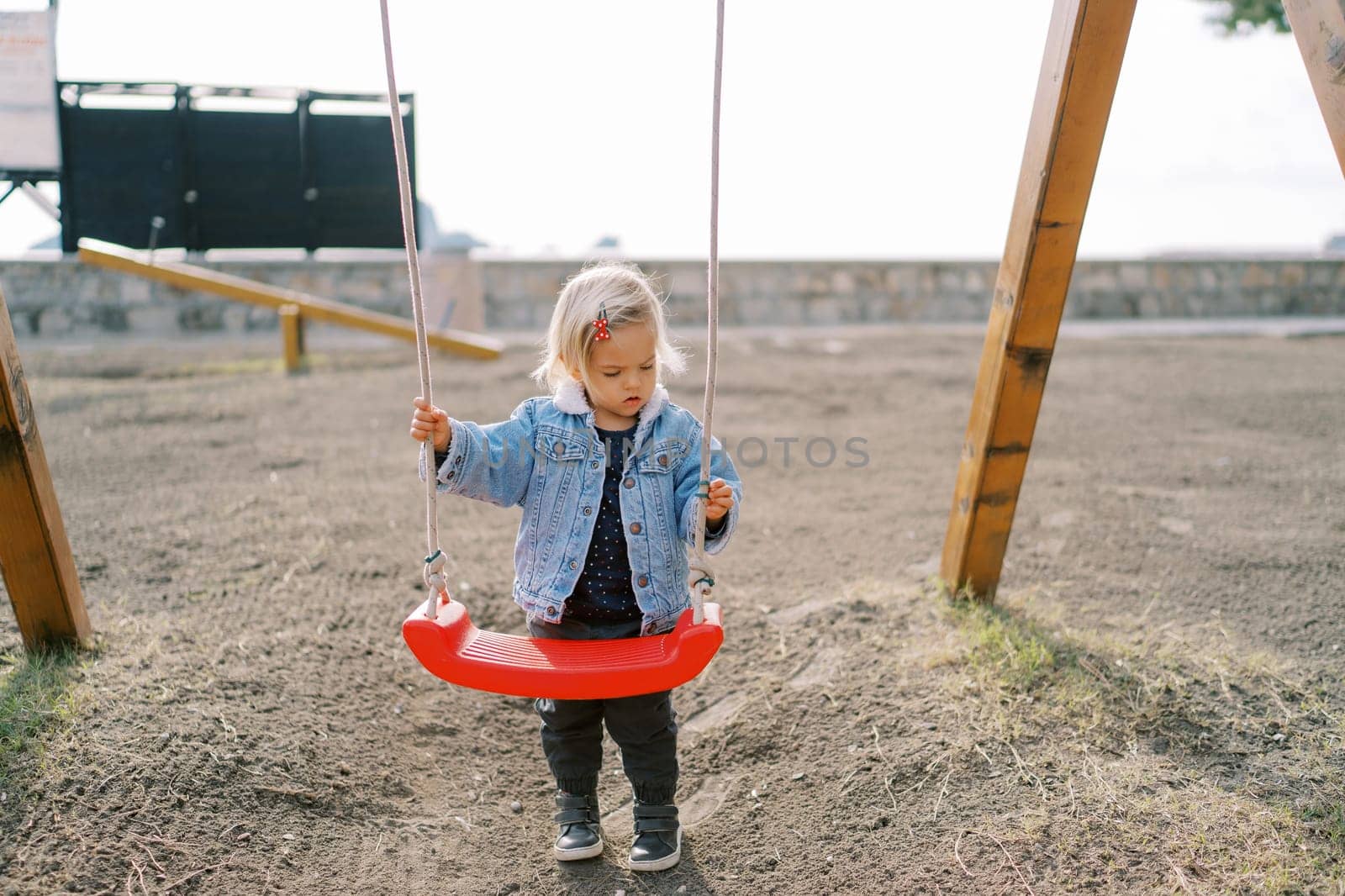 Little girl stands near the swing on the playground holding on to the ropes by Nadtochiy