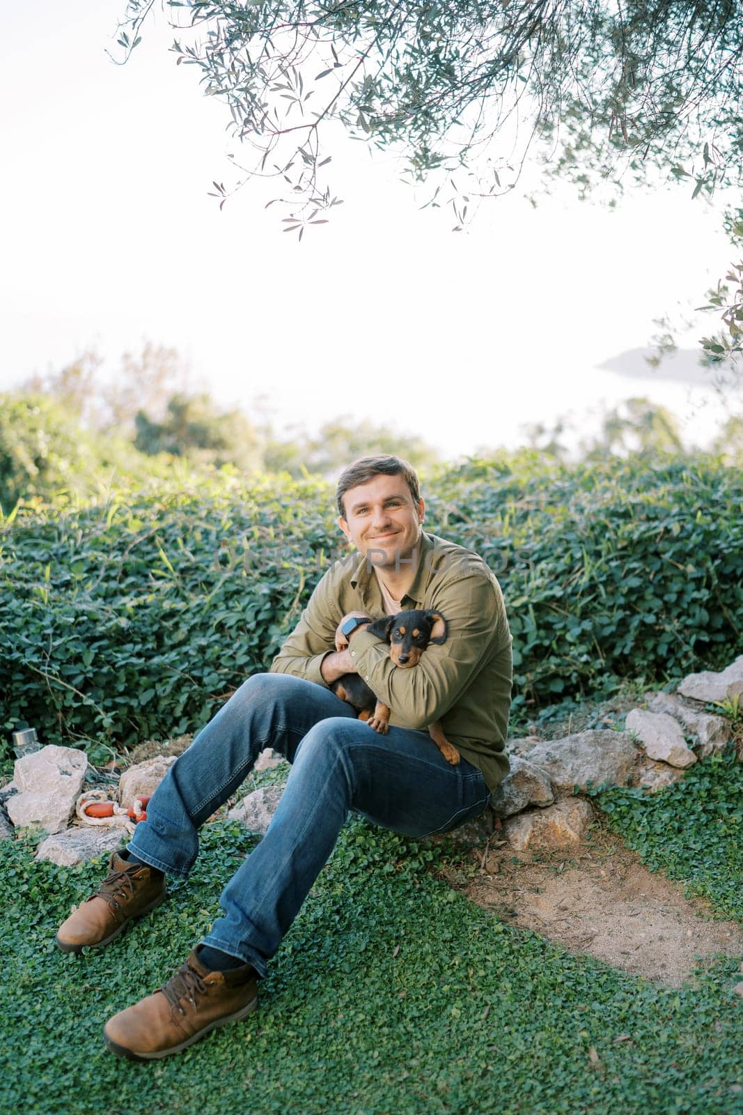 Smiling guy sitting on green grass in the park hugging a puppy on his knees. High quality photo