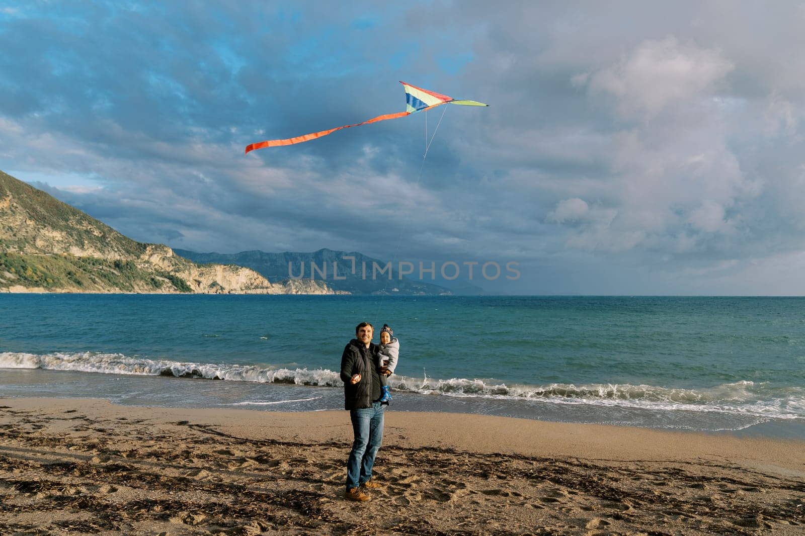 Dad with a small child in his arms stands by the sea and blows a kite by Nadtochiy