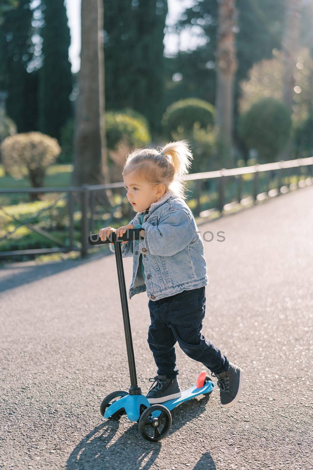 Little girl rides a three-wheeled scooter on a road in the park, pushing off with her foot. High quality photo