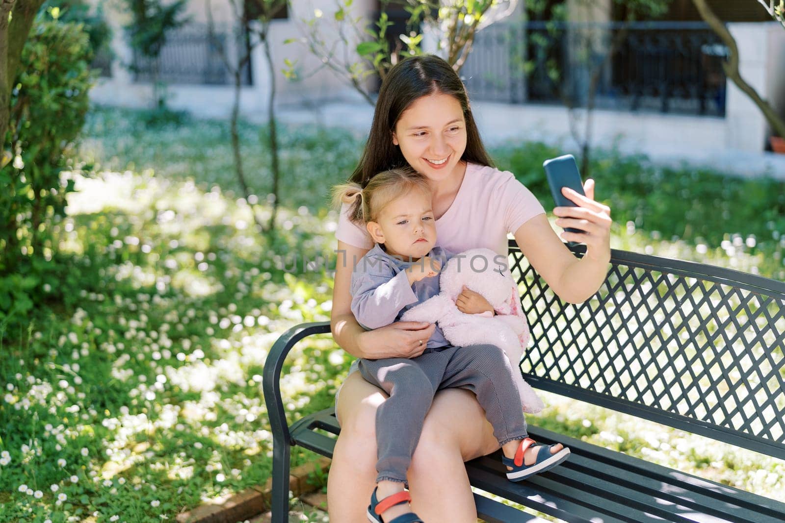 Smiling mother takes a selfie with a smartphone with a little thoughtful girl on her lap on a bench. High quality photo