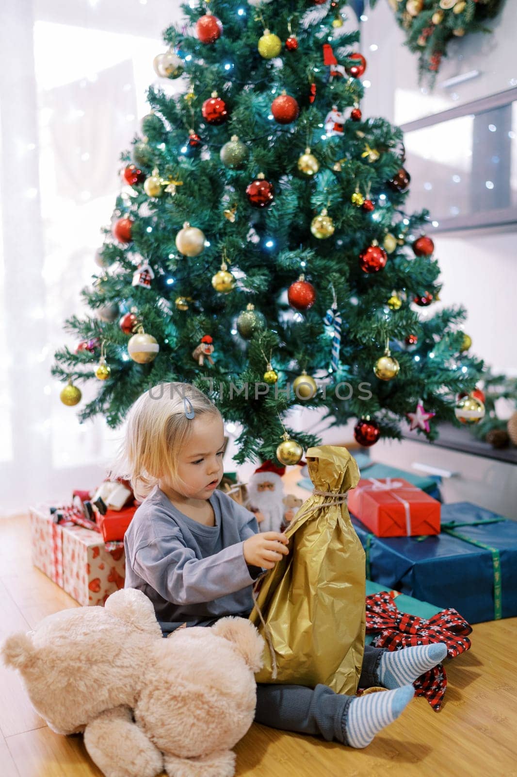 Little girl sits on the floor near the Christmas tree and unpacks a gift by Nadtochiy