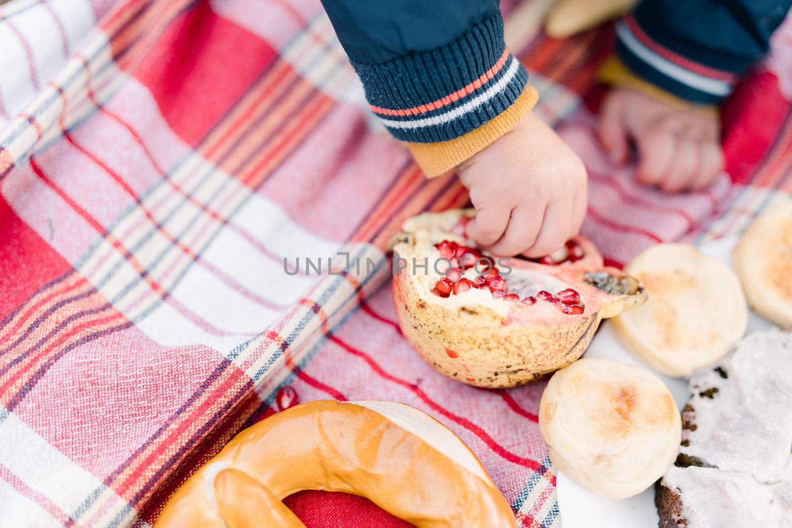 Small child picks out seeds from a pomegranate while sitting on a bedspread. Cropped. Faceless by Nadtochiy
