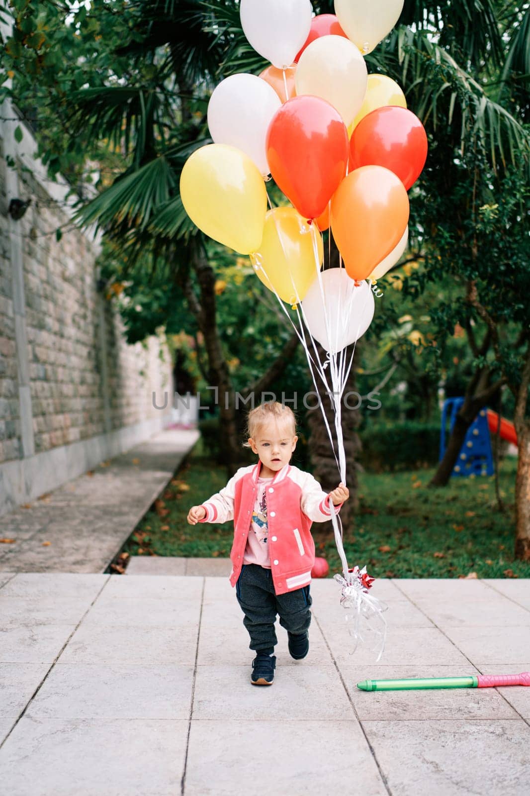 Little girl with a bunch of colorful balloons walks through a green garden by Nadtochiy