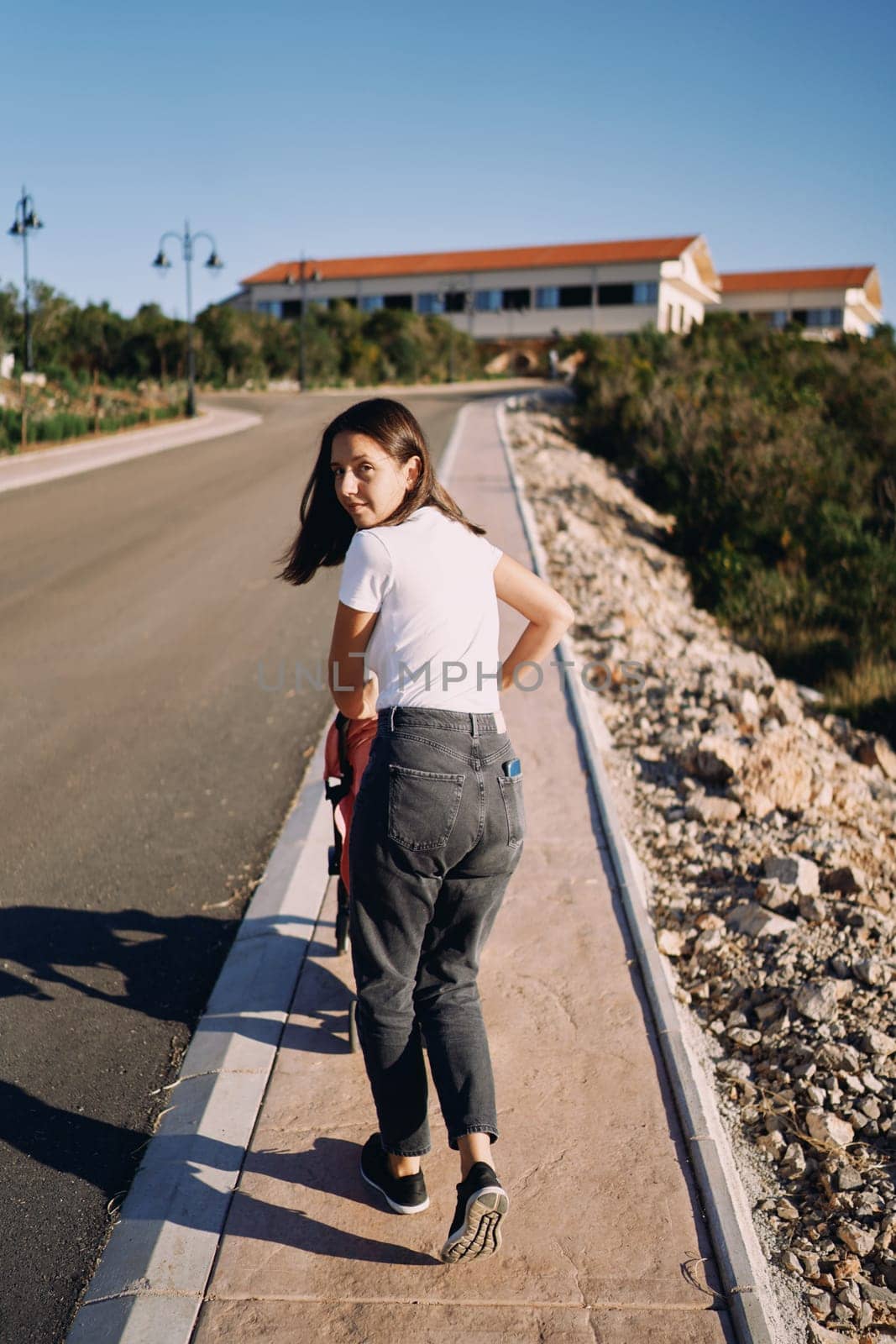 Young woman is pushing a baby carriage along the sidewalk along the road looking back. Back view by Nadtochiy