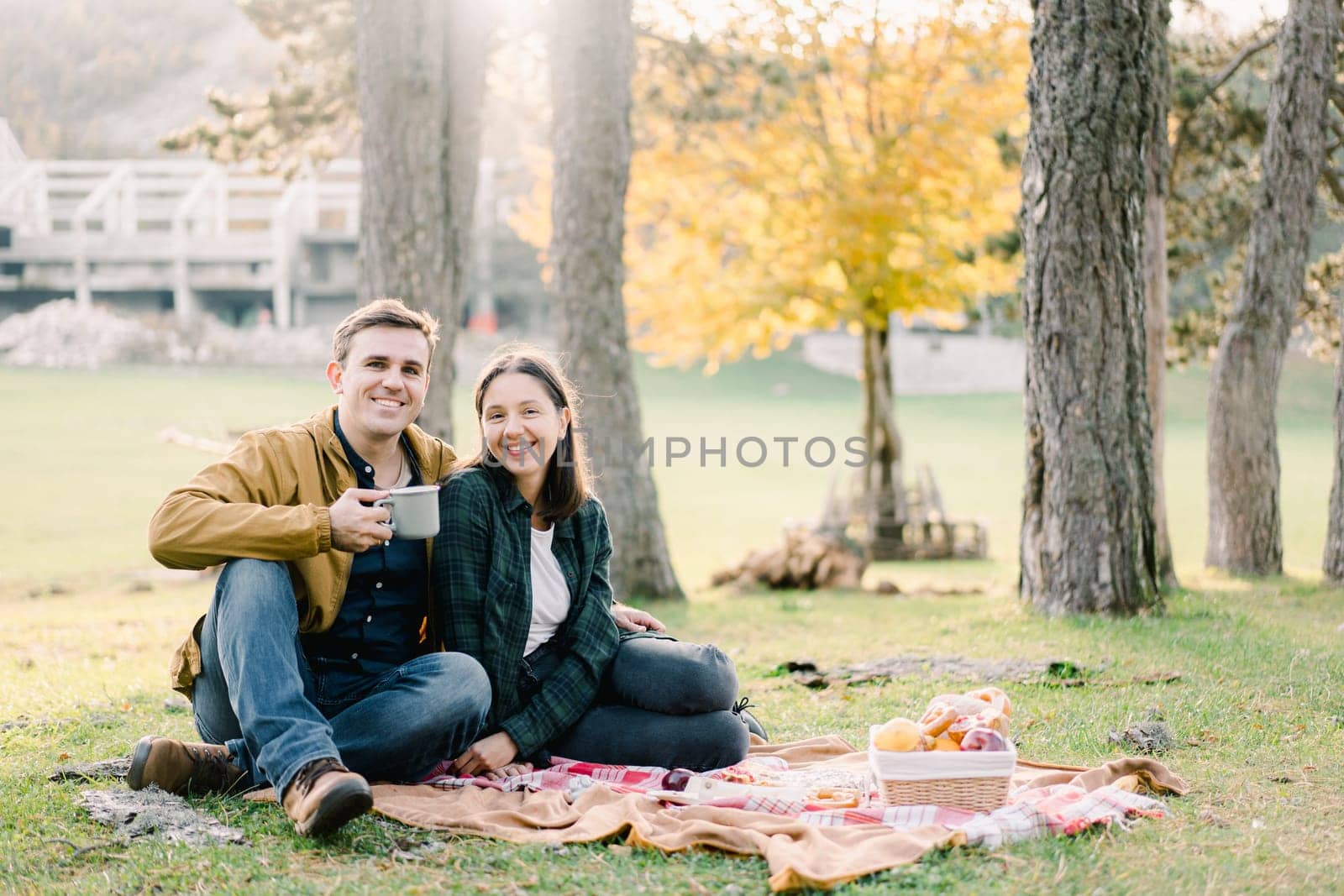 Smiling man with mug hugging woman while sitting on blanket in park near food basket by Nadtochiy