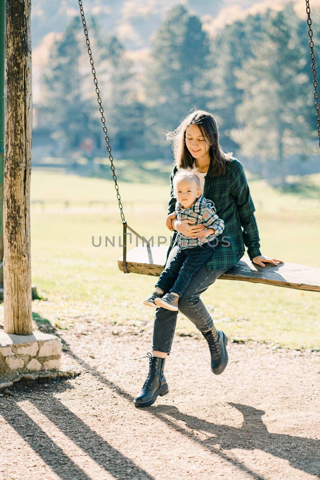 Little girl sitting on her mother lap swinging on a wooden swing in the park by Nadtochiy