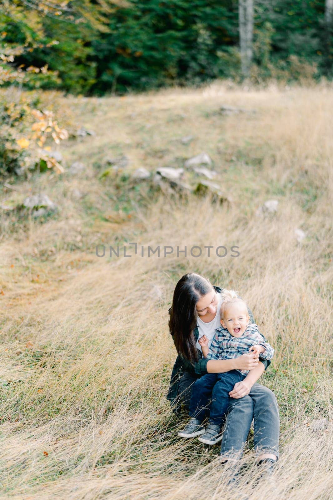 Little girl laughs while sitting in her mother arms in a clearing by Nadtochiy