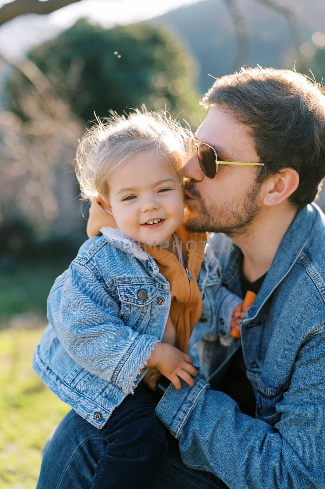 Dad kisses on the cheek a little smiling girl on his knees sitting on the lawn. High quality photo