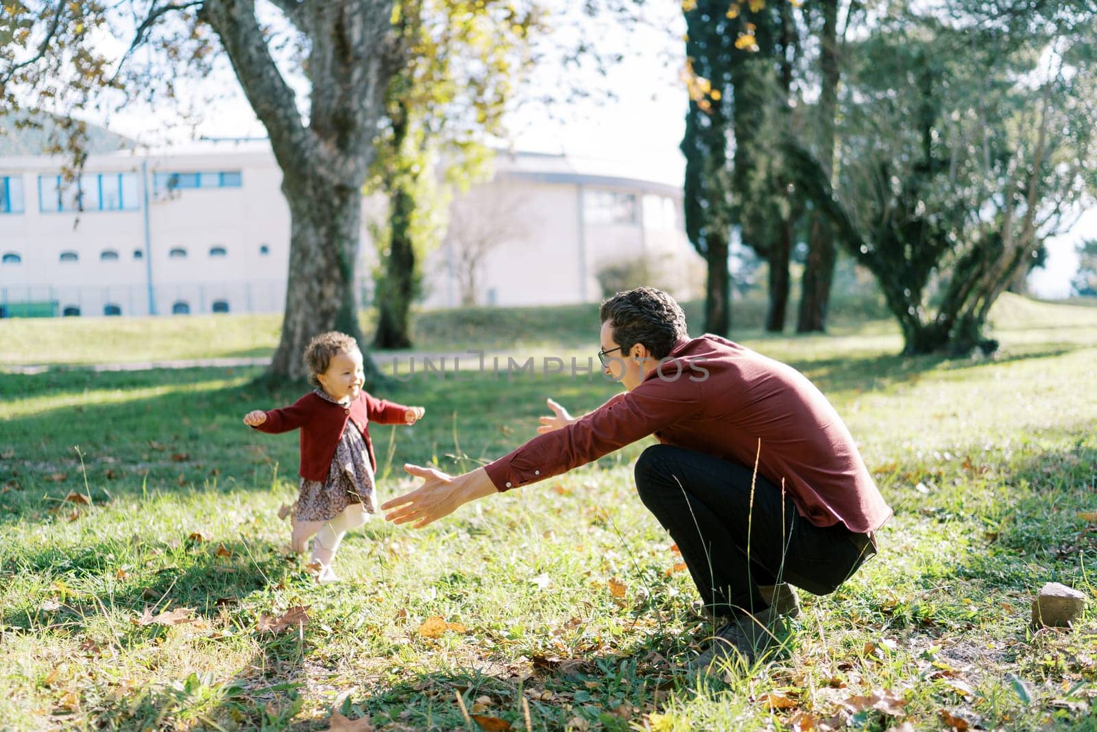 Little girl runs to her dad, who opened his arms in an embrace, squatting on the lawn. High quality photo