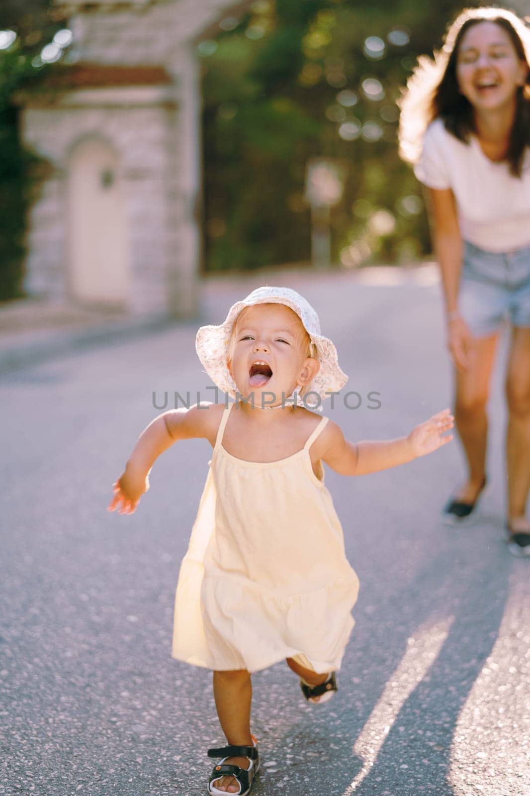 Little girl runs along the road in the park with her mouth open against the background of a laughing mother by Nadtochiy