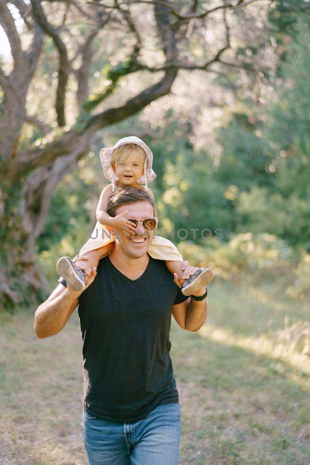 Little girl covers sunglasses on dad face with her hand, sitting on his shoulders. High quality photo