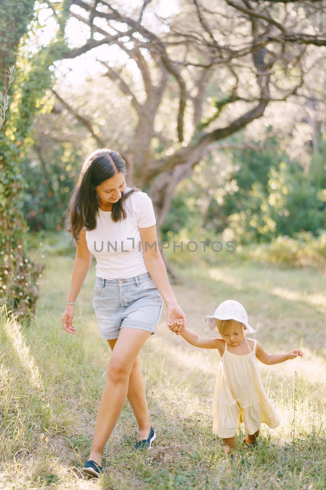 Mom leads a little girl by the hand through a green park, looking at her feet by Nadtochiy