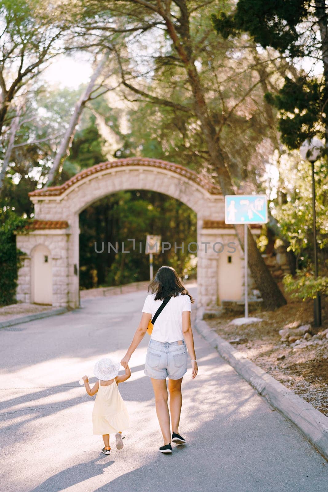 Mom leads a little girl by the hand along the road to the gates of the park. Back view. High quality photo