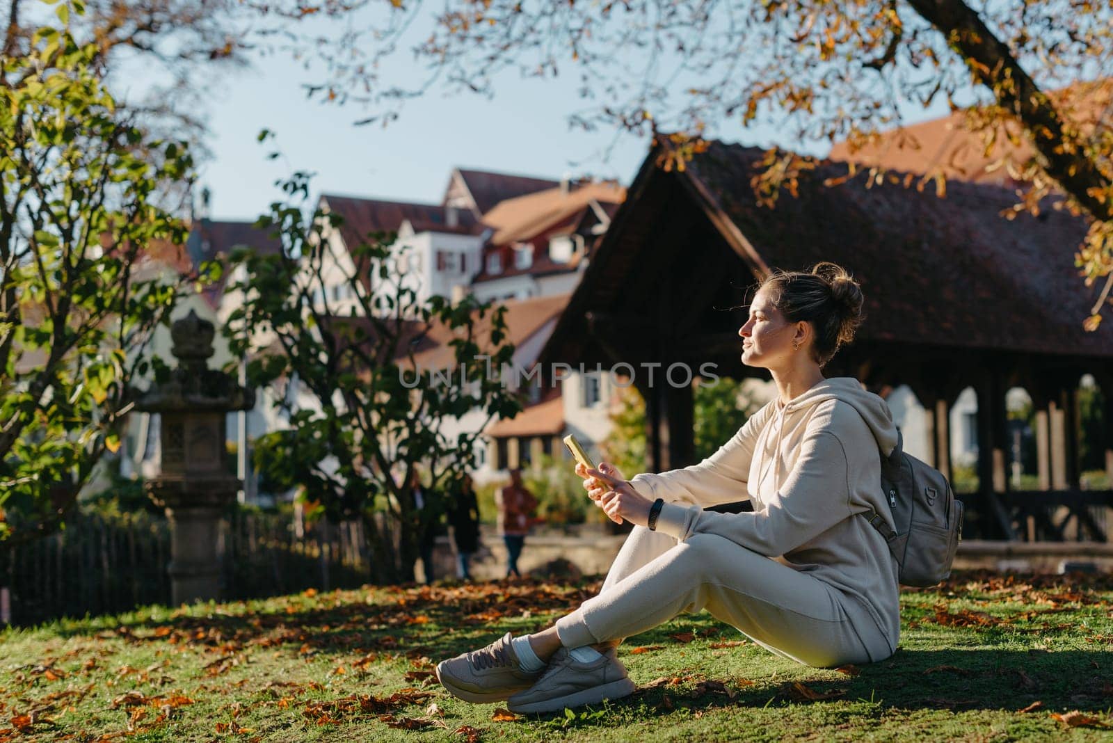 Young fashionable teenage girl with smartphone in park in autumn sitting at smiling. Trendy young woman in fall in park texting. Retouched, vibrant colors. Beautiful blonde teenage girl wearing casual modern autumn outfit sitting in park in autumn. Retouched, vibrant colors, brownish tones.