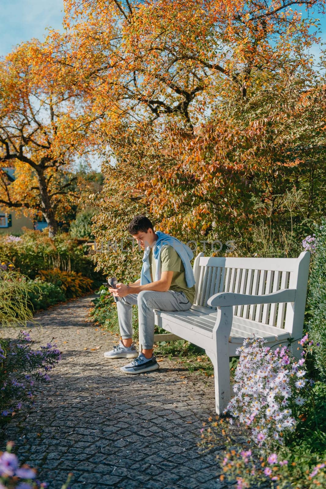 A Teenager Sits On A Bench In The Autumn Park Drinks Coffee From A Thermo Mug And Looks Into A Phone. Portrait Of Handsome Cheerful Guy Sitting On Bench Fresh Air Using Device Browsing Media Smm Drinking Latte Urban Outside Outdoor by Andrii_Ko