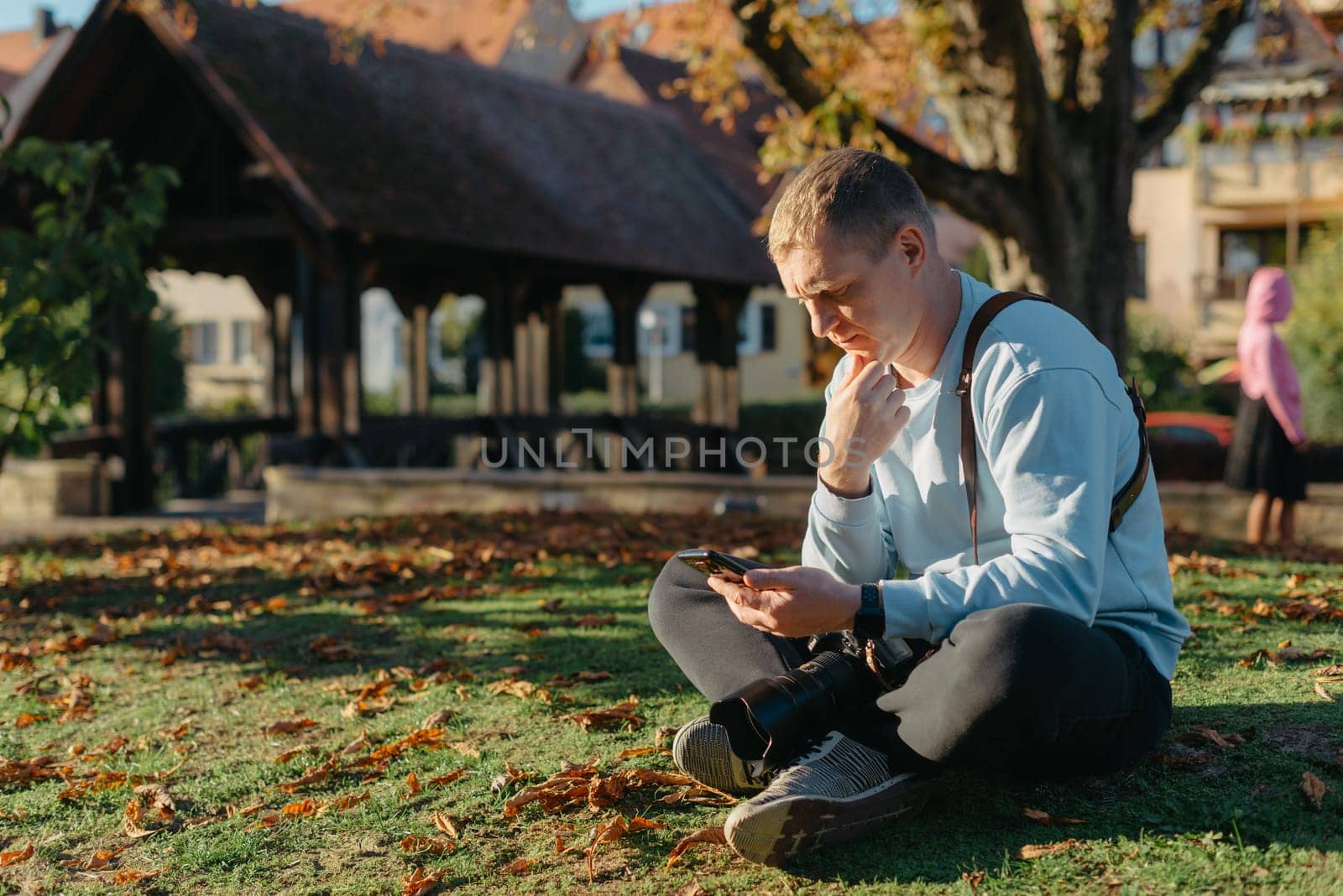 Professional Photographer Taking Picture Of Beautiful Autumn Park. Man Professional Photographer Sit With Camera And With Smartphone In Autumn Park. Retouched, Vibrant Colors, Brownish Tones. by Andrii_Ko