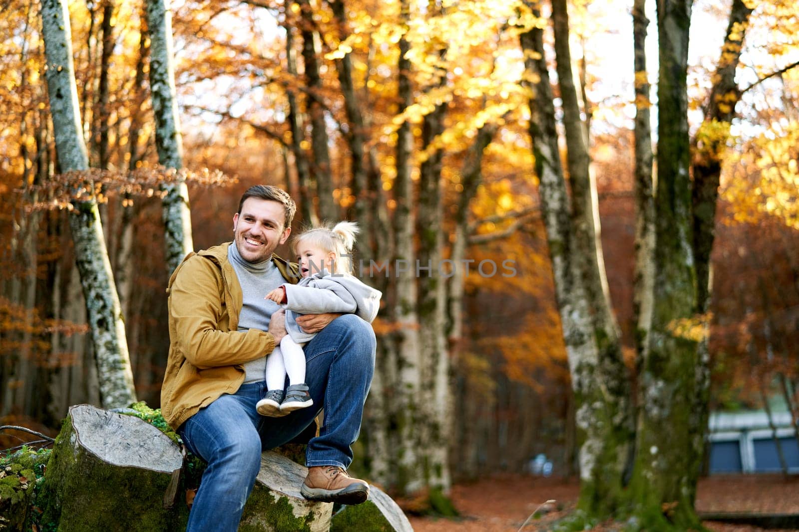 Little girl points her dad into the distance while sitting on his knees on a stump in an autumn park. High quality photo