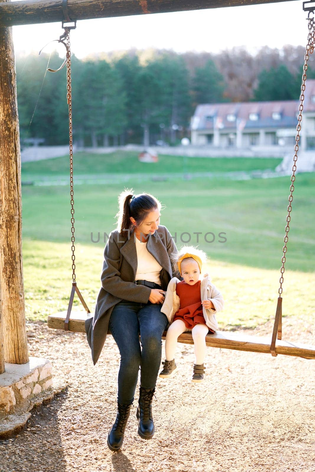 Mom hugs a little girl sitting with her on a wooden chain swing in the park by Nadtochiy