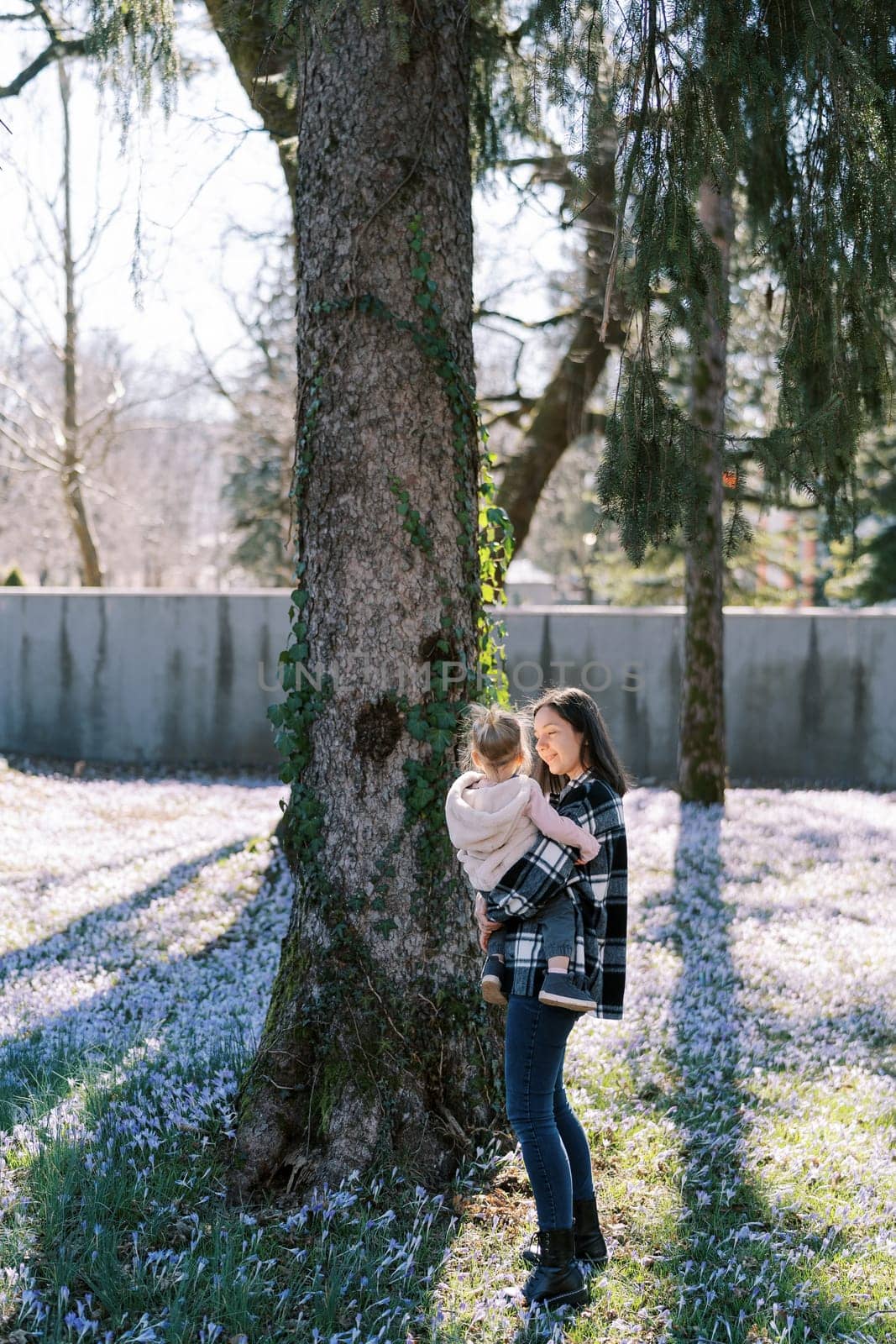 Mom with a little girl in her arms stands near an ivy-covered tree in the park by Nadtochiy