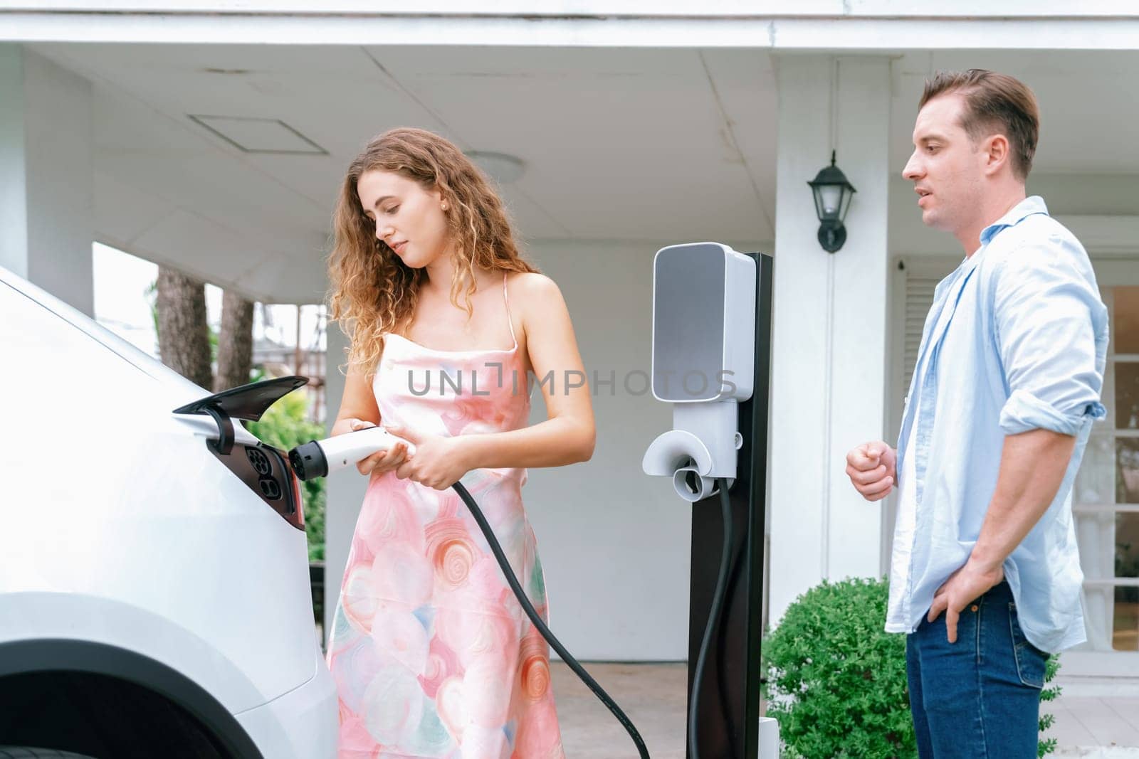 Happy and lovely couple with eco-friendly conscious recharging electric vehicle from EV home charging station. EV car technology utilized for residential home to future sustainability. Synchronos