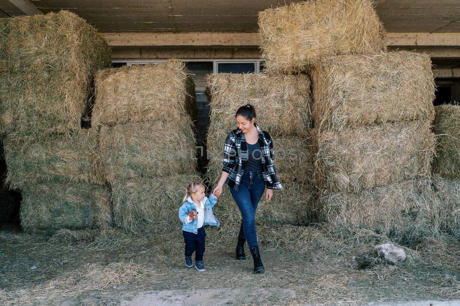 Mom and little girl walk through the hayloft past stacked bales of hay. High quality photo