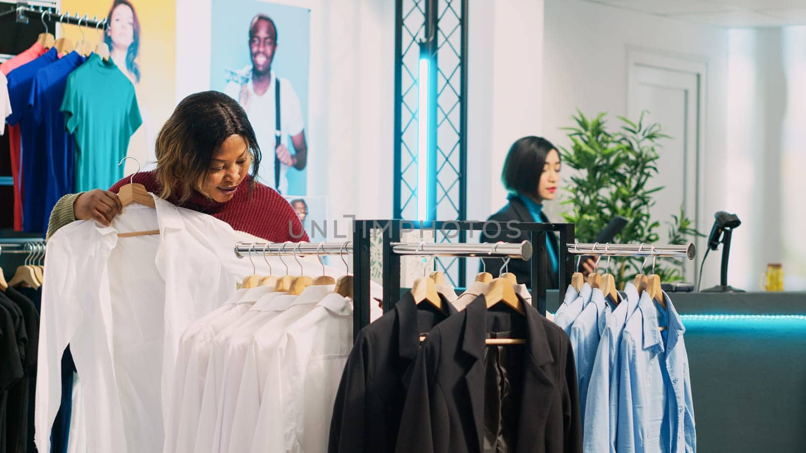 African american customer examining clothes on hangers and checking materials in clothing store. Young woman looking to buy casual or formal wear from shopping center, retail shop.