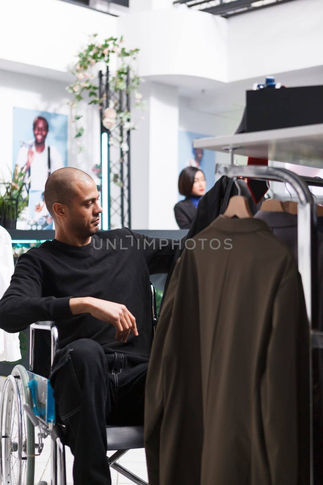 Arab man client in wheelchair choosing formal jacket and browsing through display rack in retail center showroom. Buyer with physical disability shopping for outfit in clothing store