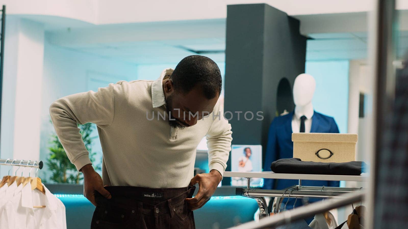 Young client checking pair of pants in clothing store, looking to buy fashionable brands on sale. Casual man examining formal wear clothes in retail shop at mall, small business. Handheld shot.