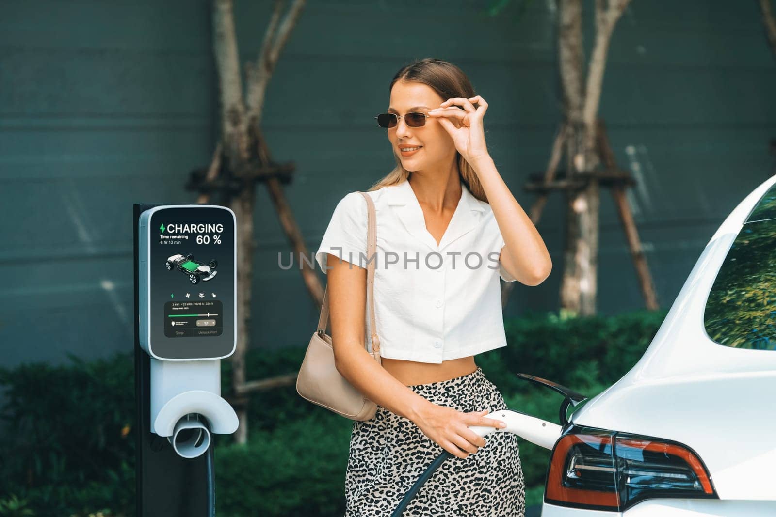 Young woman travel with EV electric car charging in green sustainable city outdoor garden in summer. Urban sustainability lifestyle by green clean rechargeable energy of electric BEV vehicle innards