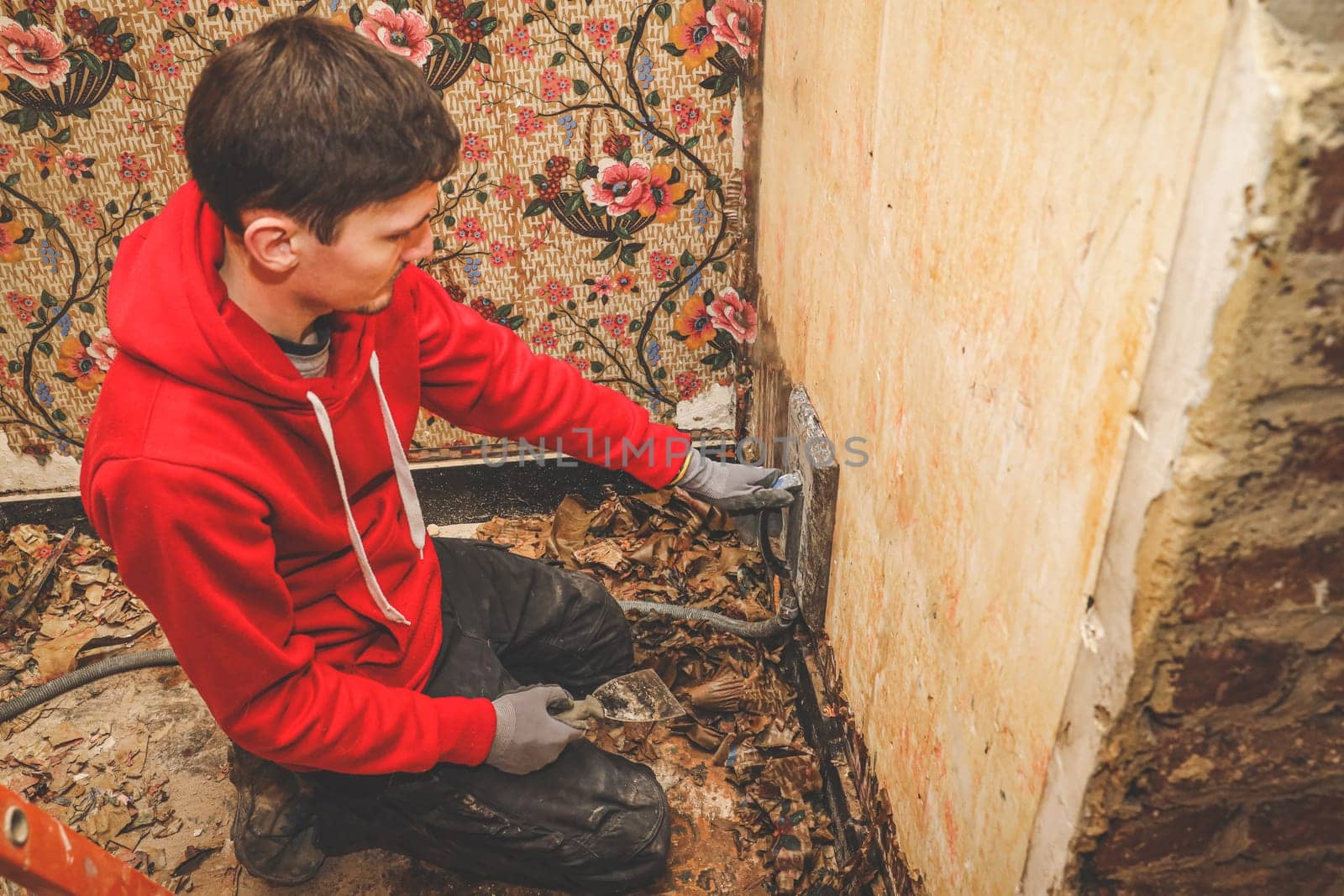A young Caucasian man in a red sweatshirt squats and removes retro wallpaper from the wall using a steam generator in an old abandoned house, close-up top view. Concept for home renovation, construction work, home.