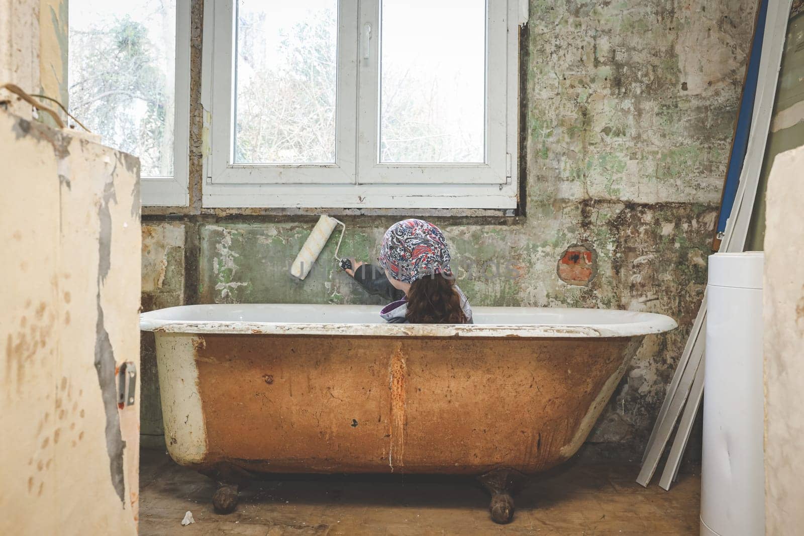 A caucasian teenage girl in a bandana sits with her back in the bathroom opposite a plastic window and holds a roller for painting walls in an old abandoned and ruined house, close-up side view.Concept home renovation, interior abandoned houses.