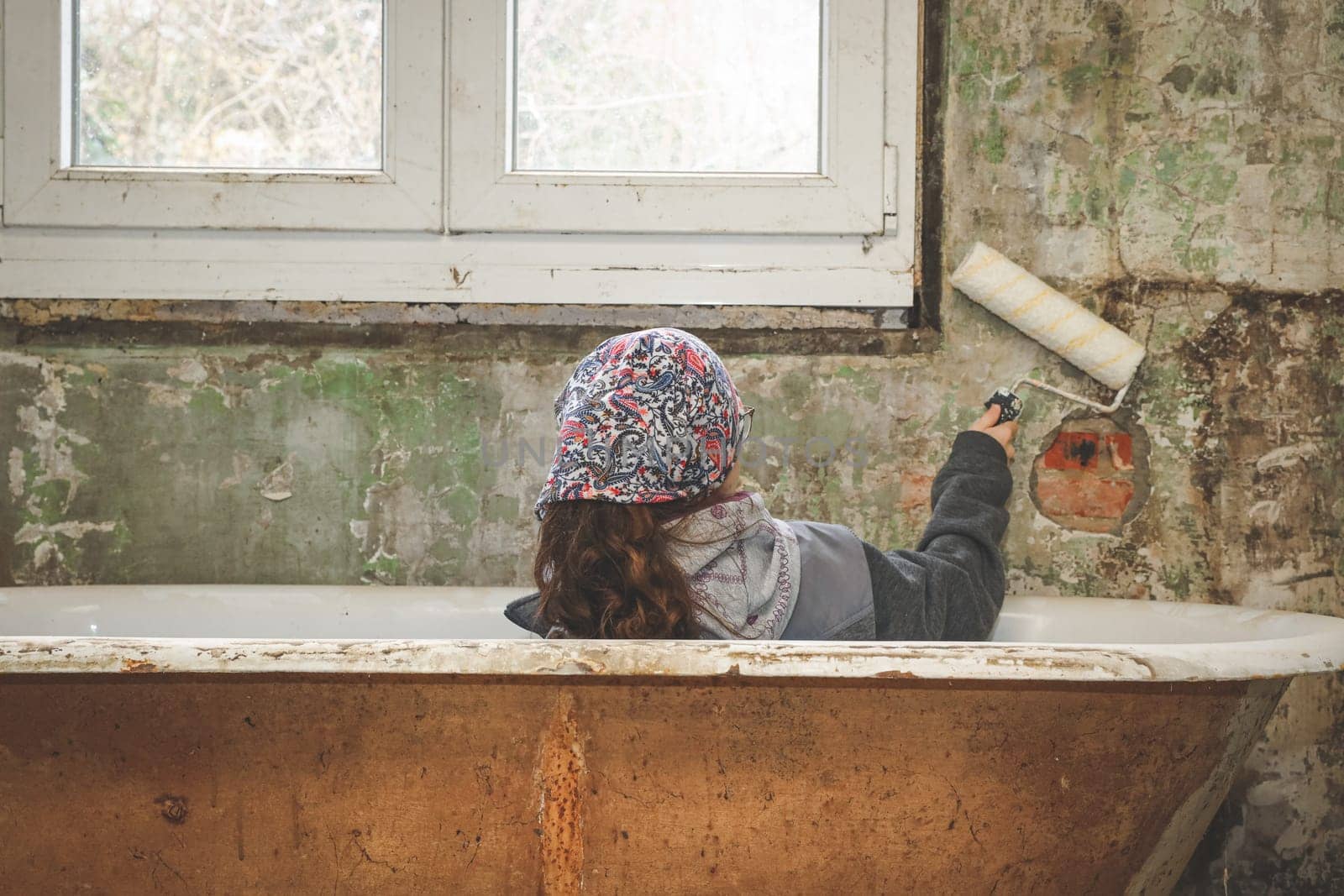 A caucasian teenage girl in a bandana sits with her back in the bathroom against a plastic window and holds a roller for painting walls in an old abandoned and ruined house, close-up side view. The concept of home renovation, the interior of abandoned houses, construction work, at home..