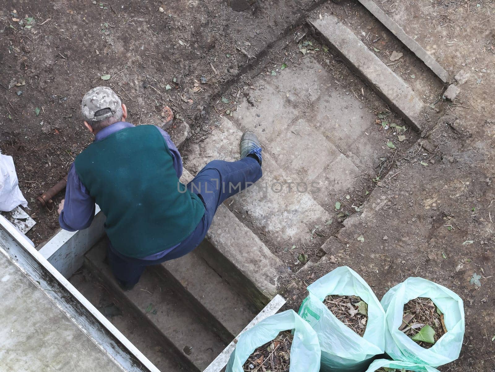 One elderly Caucasian man in casual clothes stands on stone steps in an inclined position from his back and removes garbage from the ground near the terrace in the backyard of a house on a spring day, flat lay close-up. Concept for home renovation, construction work, home.