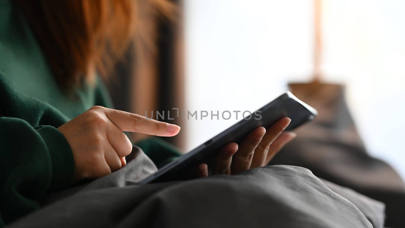 Young woman in green sweater browsing internet or checking social media on digital tablet at home by prathanchorruangsak