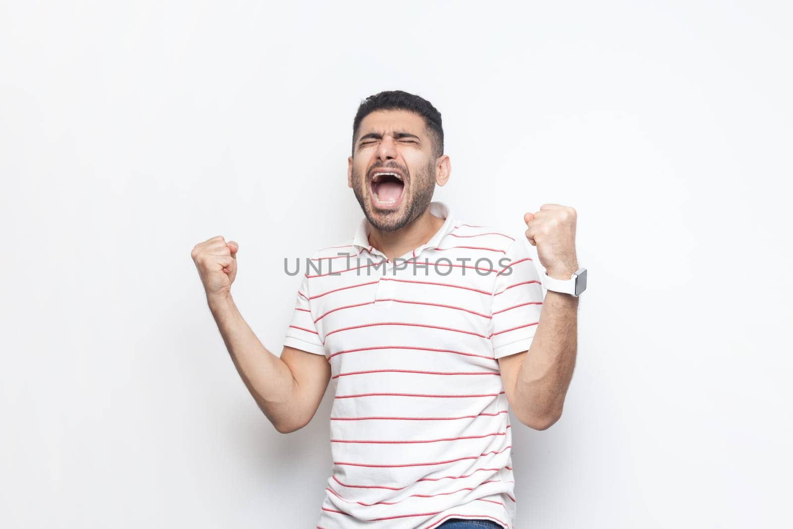 Portrait of extremely happy joyful cheerful bearded man wearing striped t-shirt standing with clenched raised fists, screaming, celebrating victory. Indoor studio shot isolated on gray background.