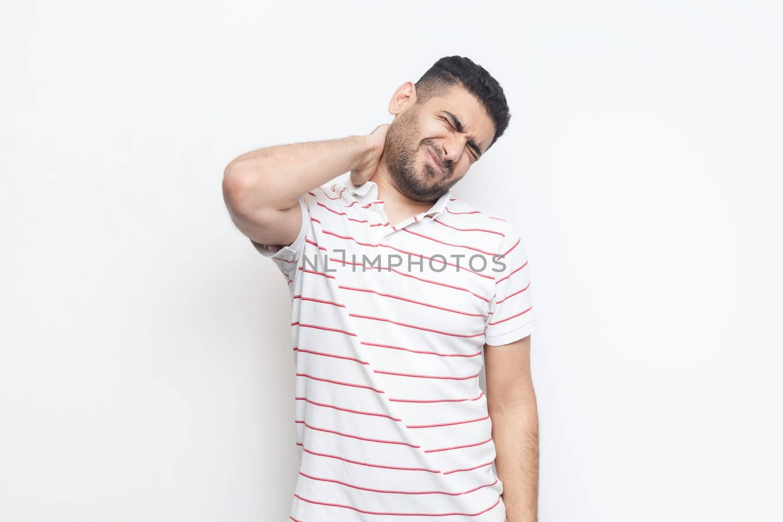 Portrait of handsome sick tired bearded man wearing striped t-shirt standing massaging his painful neck, frowning face from terrible pain. Indoor studio shot isolated on gray background.