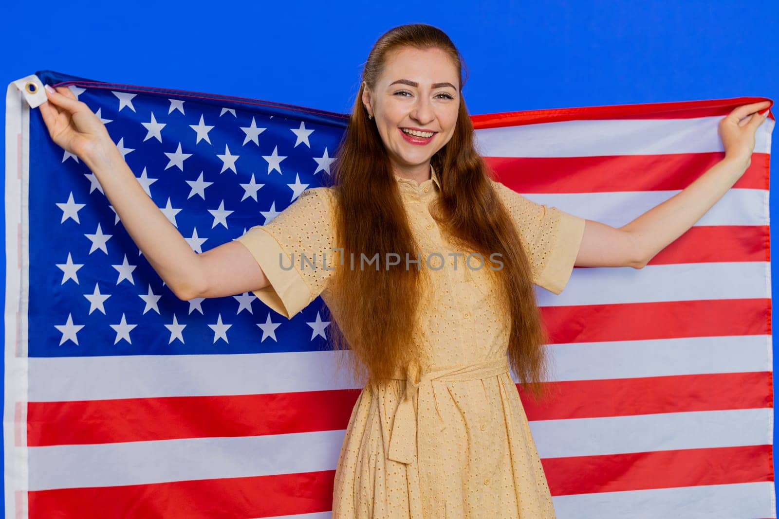 Young adult woman waving and wrapping in American USA flag, celebrating, human rights and freedoms. Independence day. Pretty attractive caucasian girl isolated on blue studio background indoors