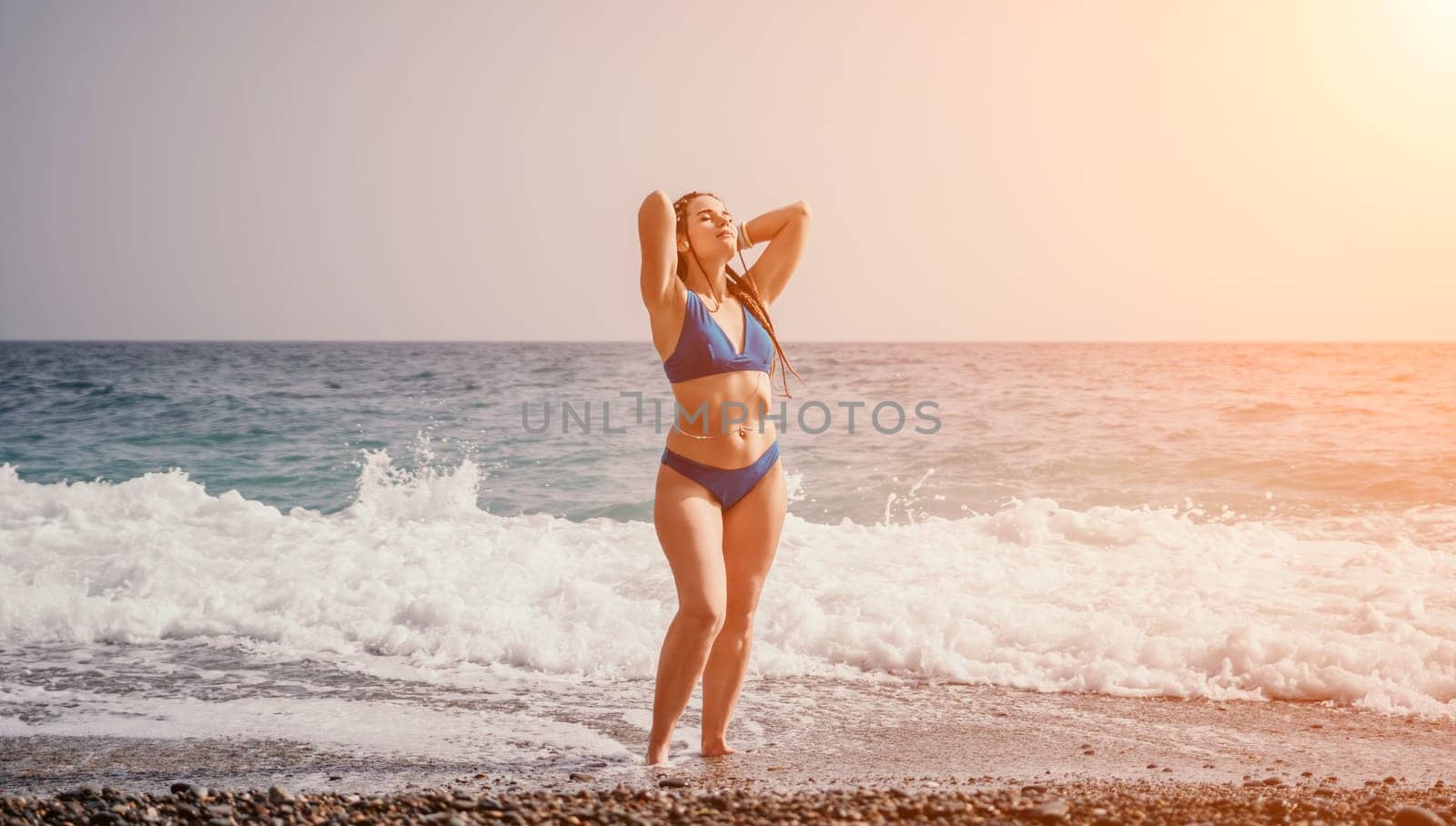 Beach vacation. Hot beautiful woman in sunhat and bikini standing with her arms raised to her head enjoying looking view of beach ocean on hot summer day.