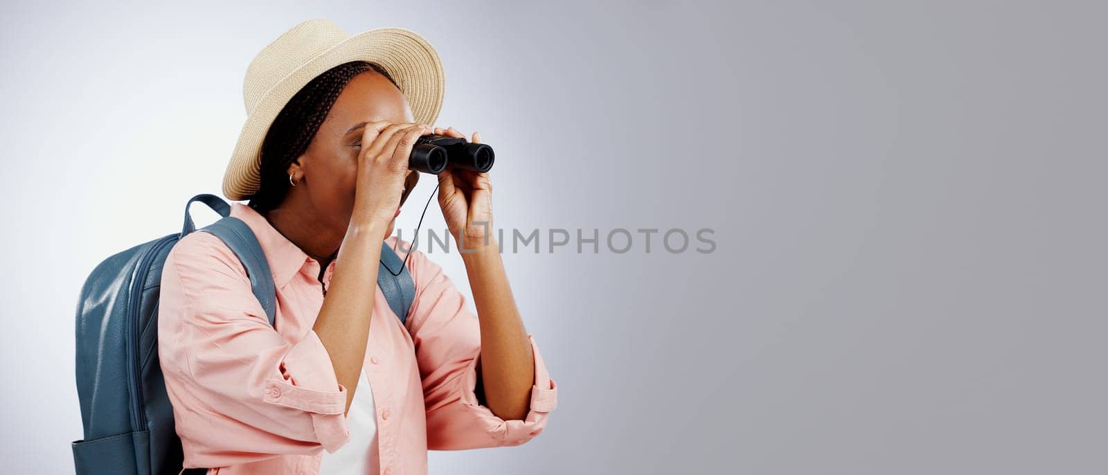 Woman, binoculars and explore on adventure, studio and travel or backpack by gray background. Black person, vacation and holiday in mockup, watch and search on journey, gear and equipment for tourist by YuriArcurs