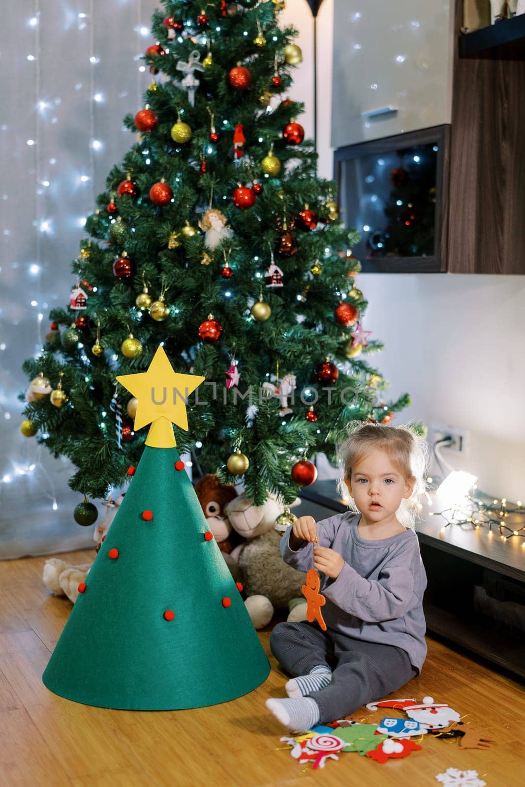 Little girl holds a felt toy in her hands while sitting on the floor near a toy Christmas tree. High quality photo