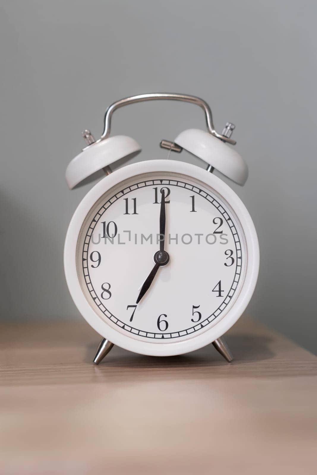 Close-up of a round white alarm clock on a table in the bedroom. The hands on the clock show seven o'clock in the morning, time to get up. Retro alarm clock on the table, vintage tone. space for text