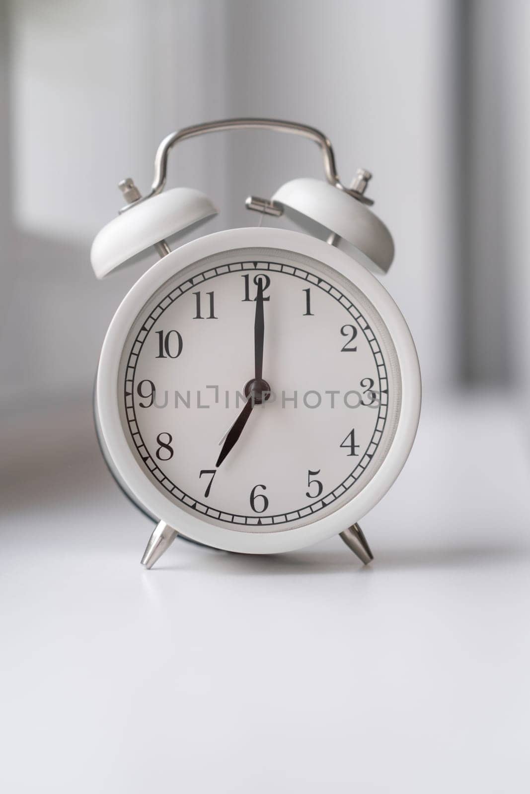 Close-up of a round white alarm clock on the windowsill in the bedroom. The hands on the clock show seven o'clock in the morning, time to get up. Retro alarm clock on the table. space for text