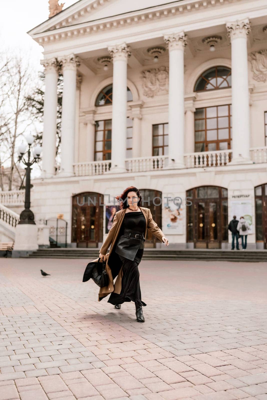 Woman street lifestyle. Image of stylish woman walking through European city on sunny day. Pretty woman with dark flowing hair, dressed in a beige raincoat and black, walks along the building